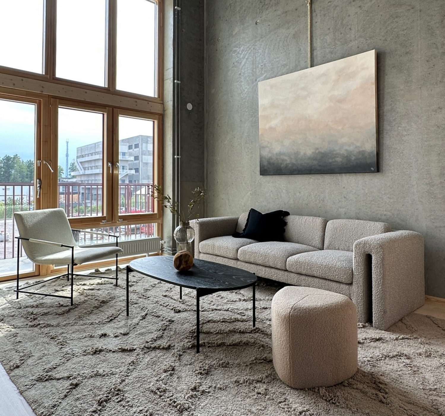Ett stilfullt inrett vardagsrum med betongväggar, stora fönster och neutrala möbler. På väggen hänger en canvastavla med en gradient från beige till grå