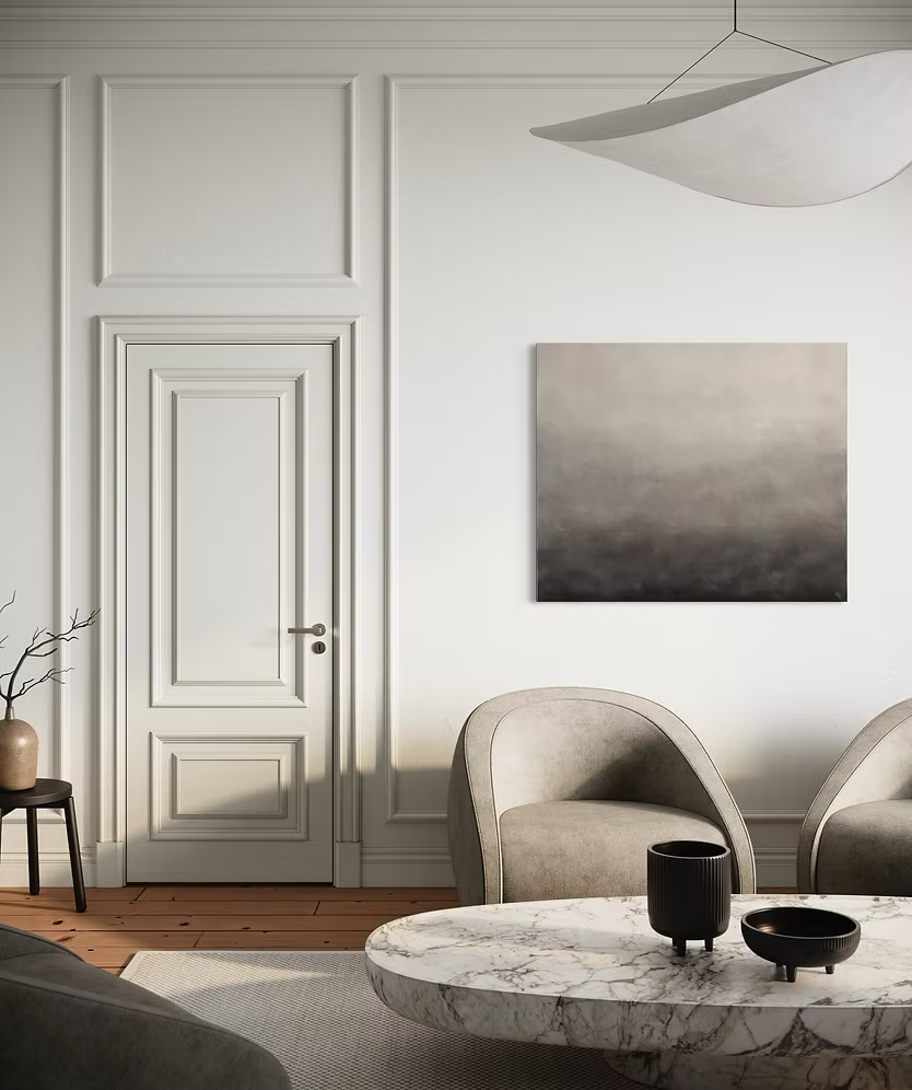 Stillfullt vardagsrum med två beige fåtöljer som står bakom ett soffbord i vit marmor. Ovanför fåtöljerna, intill en vit dörr, hänger en abstrakt målning med mjuka skiftningar i greige